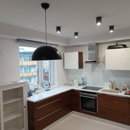 Piotr Wiktorowski - Nowoczesna kuchnia z drewnianymi szafkami, białymi blatami i wiszącą lampą. Widoczne okno z widokiem na budynek. Minimalistyczny design i jasne wnętrze.