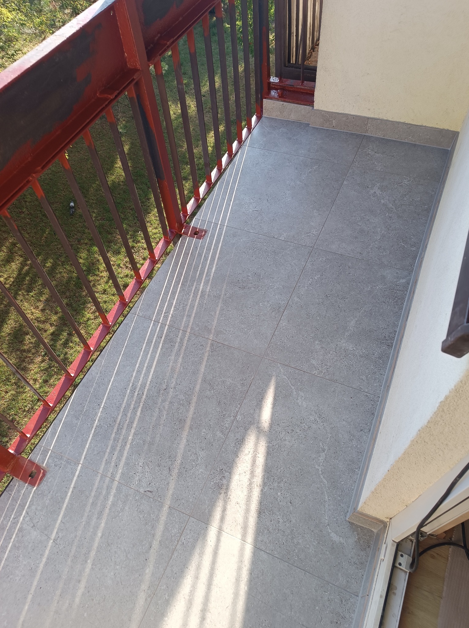 Szare płytki gresowe na balkonie z czerwoną barierką. Widoczne cienie rzucane przez barierki. Minimalistyczny design i staranne ułożenie płytek.