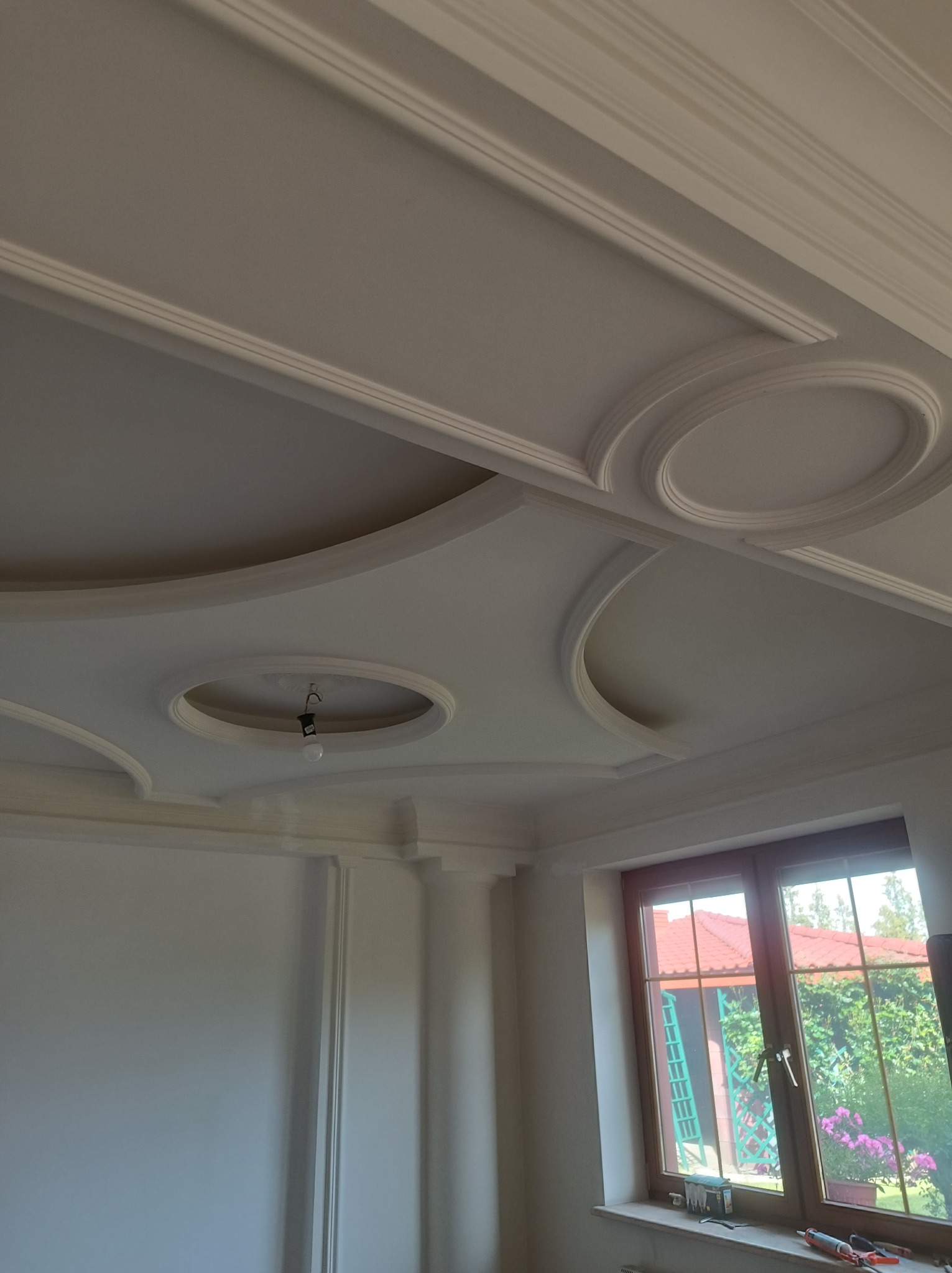 Dekoracyjny sufit z gładzią i sztukaterią w jasnym wnętrzu. Widoczne zdobienia sufitowe, okno z widokiem na zieleń i fragment ściany z elementami dekoracyjnymi.