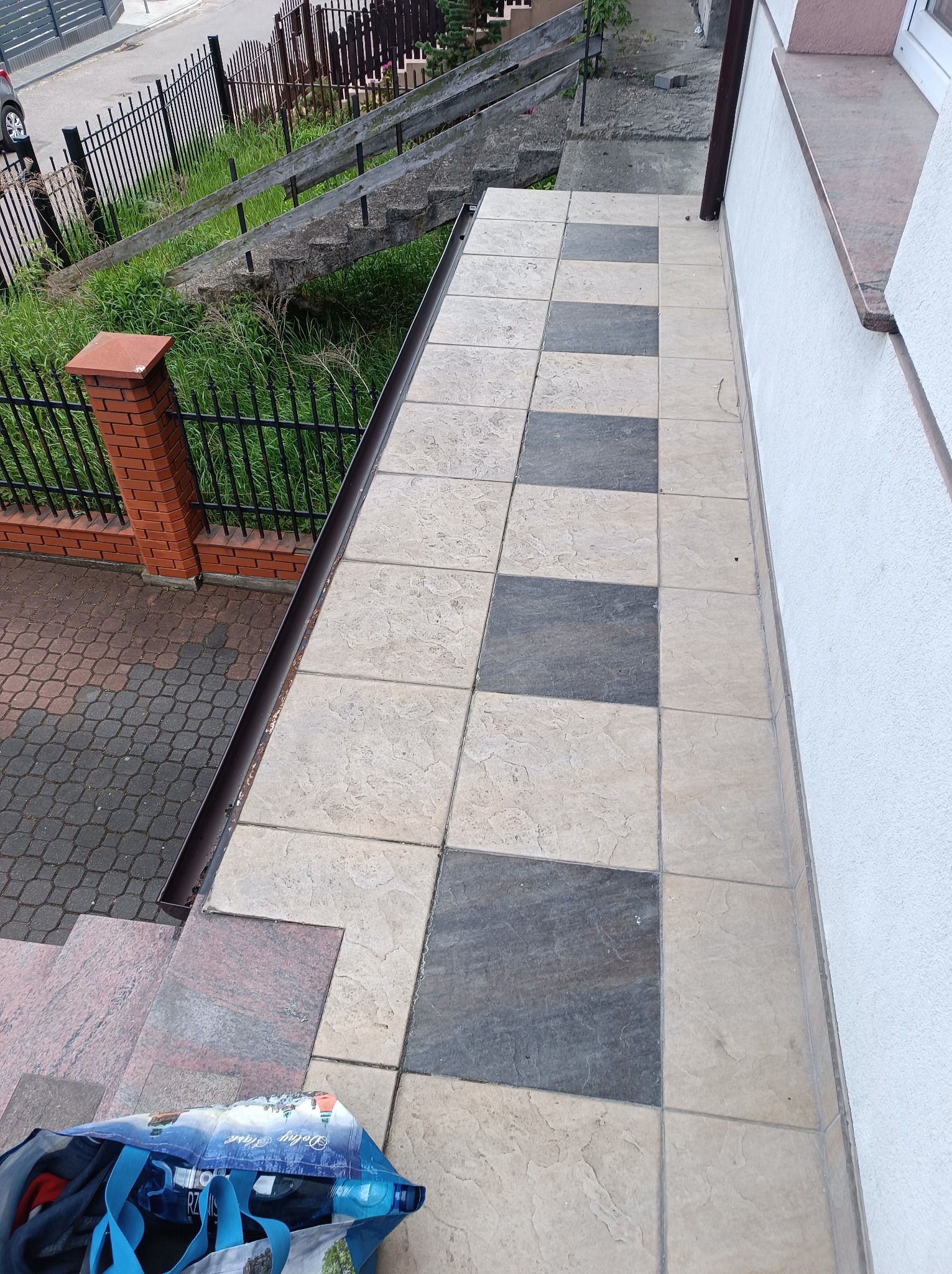 Ułożone płytki na zewnątrz budynku w szachownicę, widok z góry. Płytki w odcieniach beżu i szarości. Widoczny fragment elewacji i schodów.