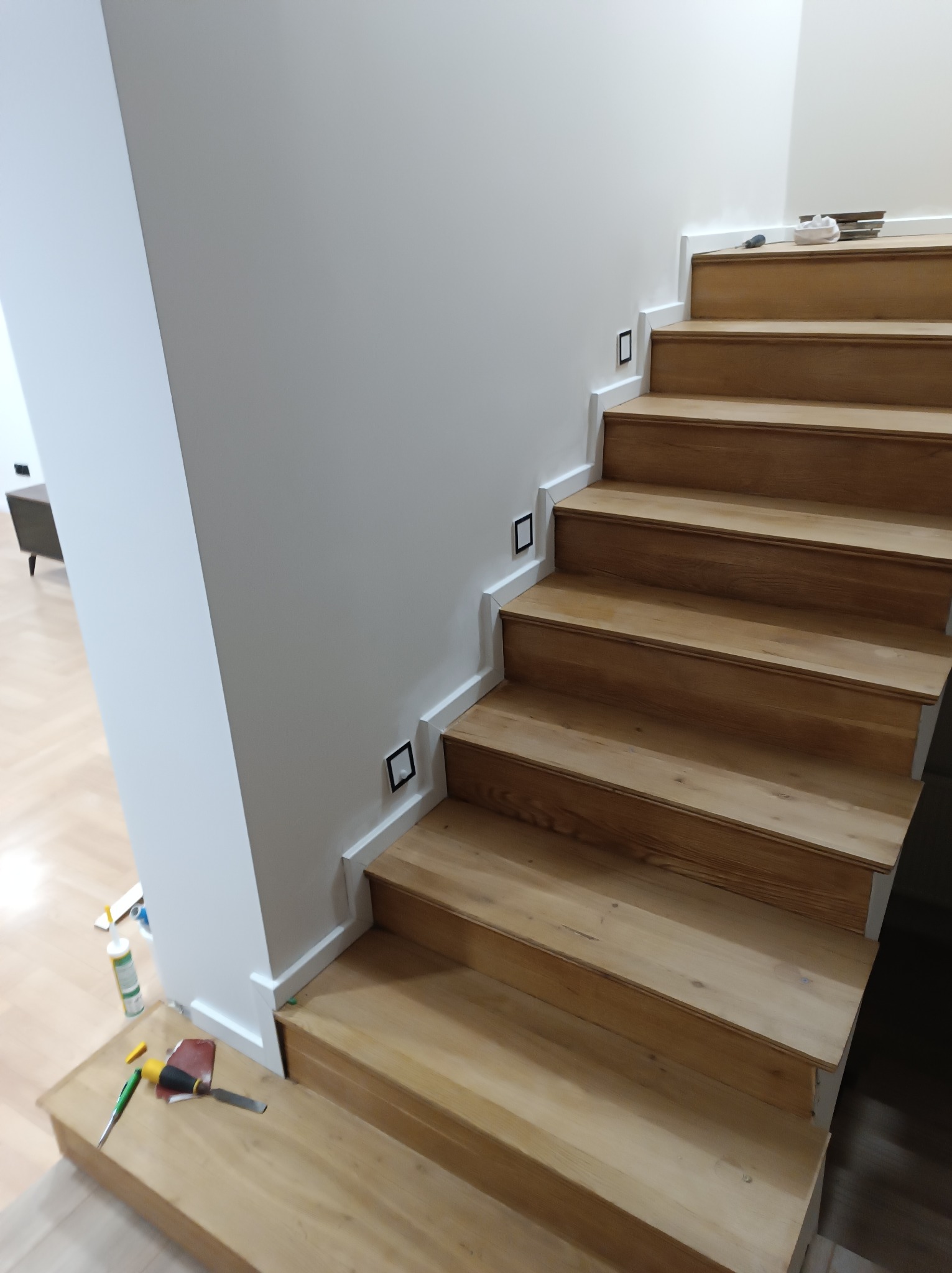 Schody drewniane z oświetleniem punktowym przy ścianie, wykończone białą listwą. Widoczne narzędzia i materiały na jednym ze stopni. Ujęcie z dołu.