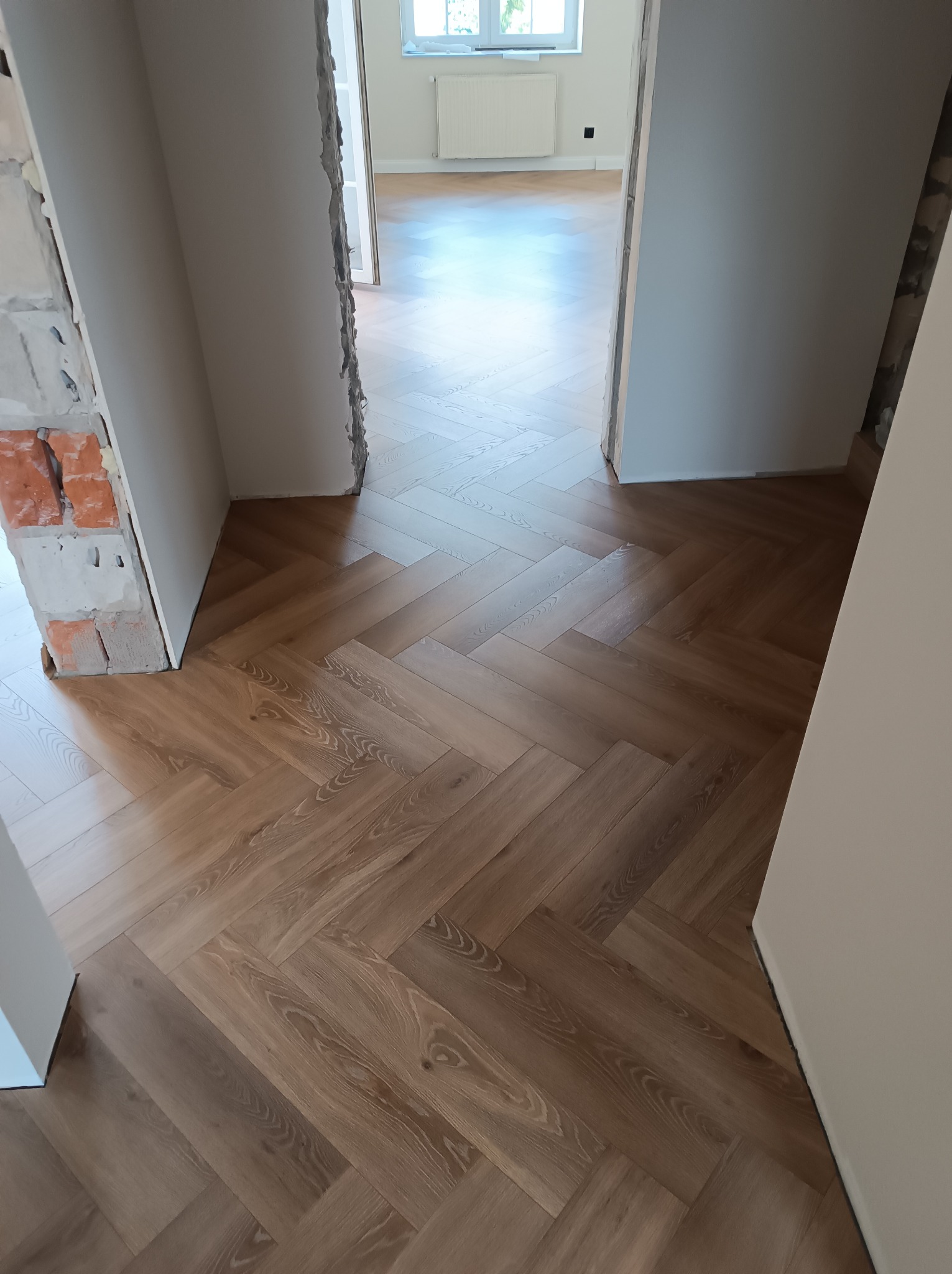 Ułożona podłoga z paneli w jodełkę w odcieniach brązu, widok z perspektywy przejścia między pomieszczeniami, surowy mur z cegieł po lewej stronie.