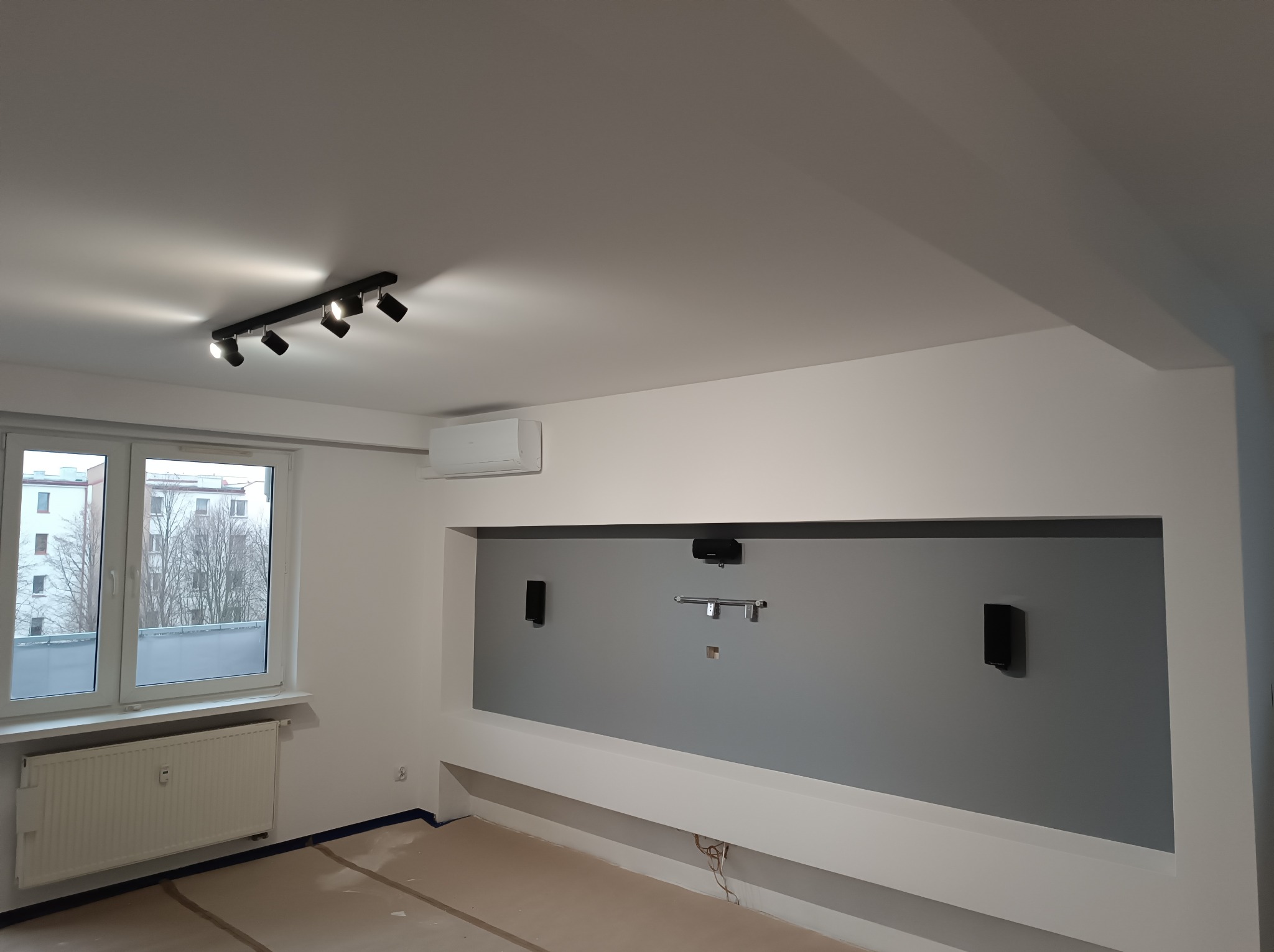 Szare wnętrze pokoju z oknem, grzejnikiem i przygotowaną ścianą pod sprzęt audio-video, oświetlenie sufitowe. Widoczne zabezpieczenia podłogi.