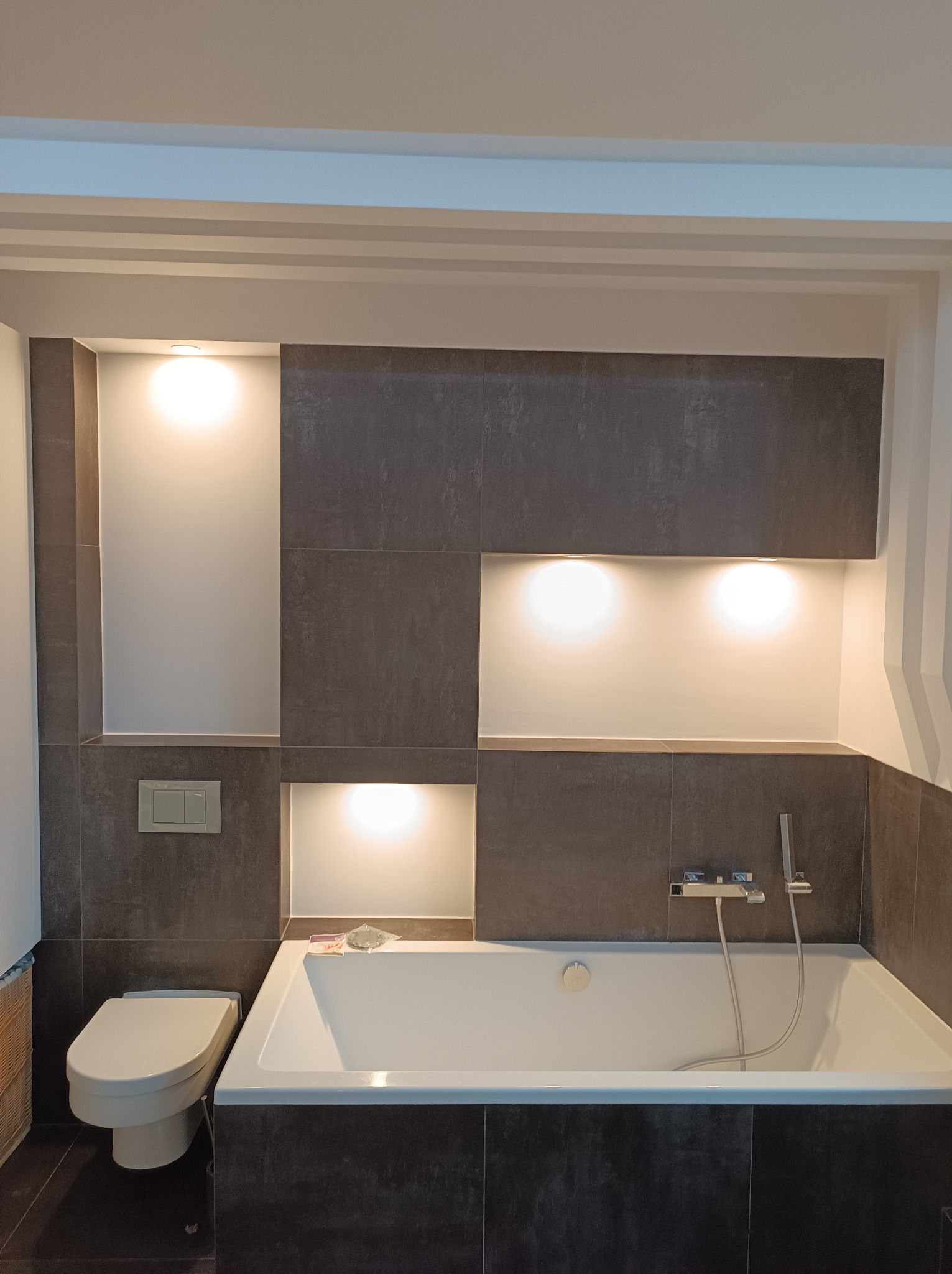 Nowoczesna łazienka z wanną, szarą okładziną i podświetlanymi wnękami. Minimalistyczny design, armatura chromowana, widoczny fragment toalety.