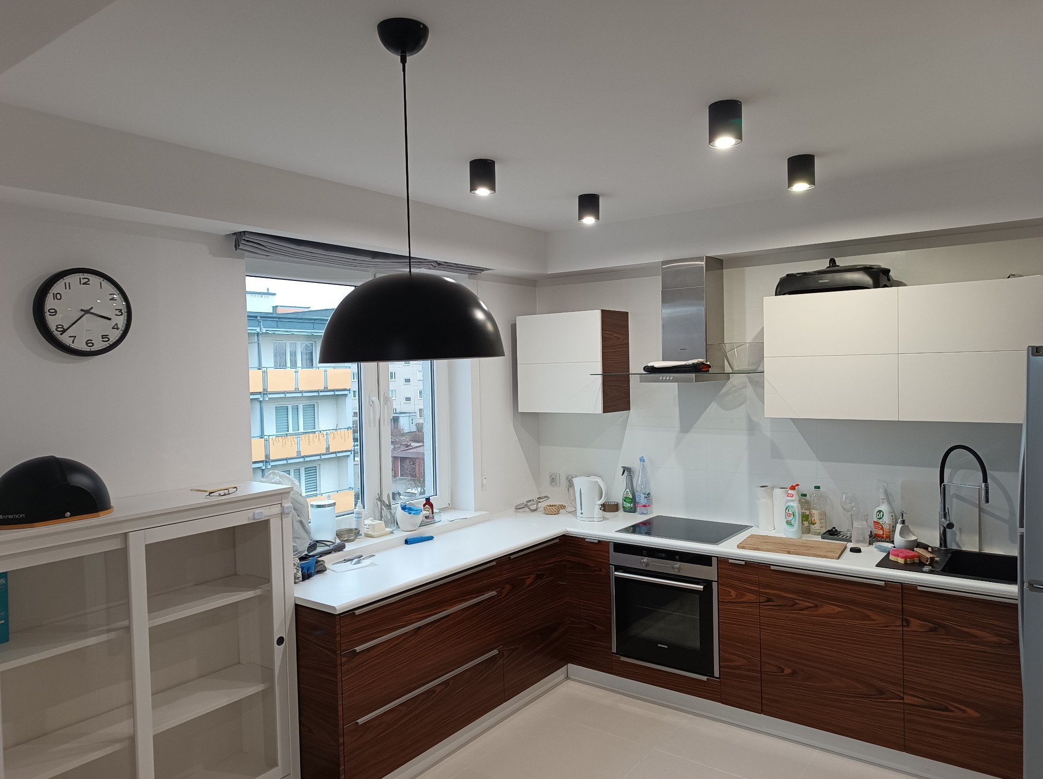 Nowoczesna kuchnia z drewnianymi szafkami, białymi blatami i wiszącą lampą. Widoczne okno z widokiem na budynek. Minimalistyczny design i jasne wnętrze.