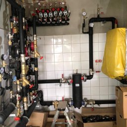 Rafał Jarząbek Rafinstal - Rozbudowana instalacja hydrauliczna i grzewcza z licznymi rurami, zaworami i manometrami, zamontowana na ścianie wyłożonej białymi płytkami, z drabinką i kartonami na podłodze.