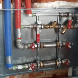 Rozdzielacz instalacji wodnej w metalowej obudowie, z rurami w izolacji czerwonej i niebieskiej, zamontowany na tle ceglanej ściany z pianką uszczelniającą.