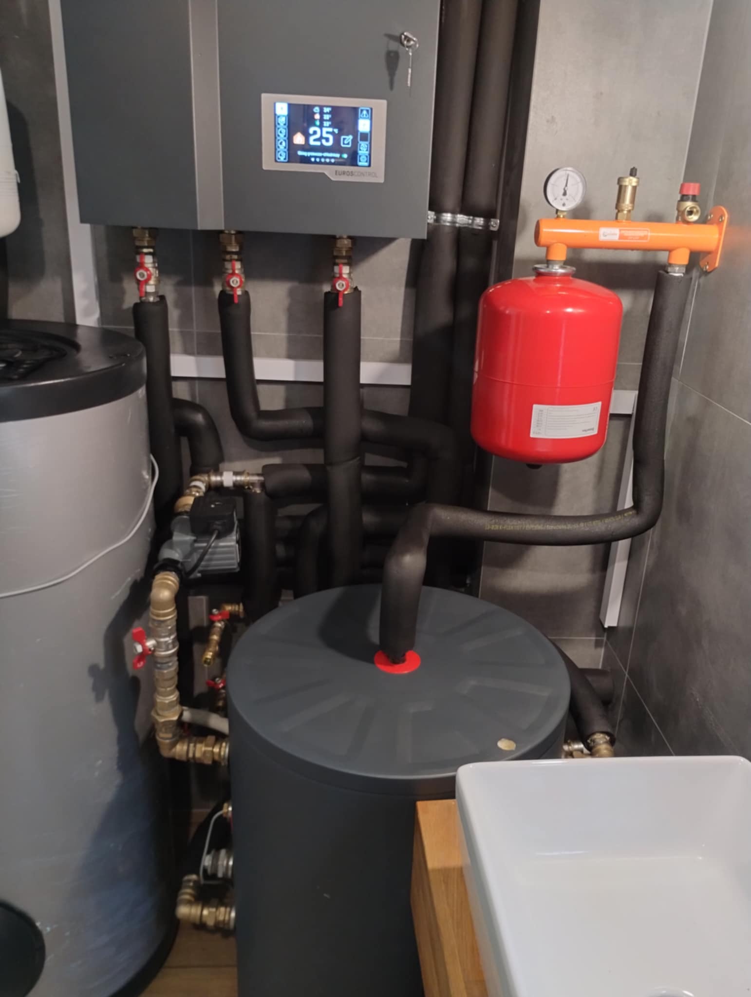 Szara kotłownia z czarnymi rurami, czerwonym zbiornikiem wyrównawczym i panelem sterowania z wyświetlaczem, widoczna w łazience z szarymi płytkami.