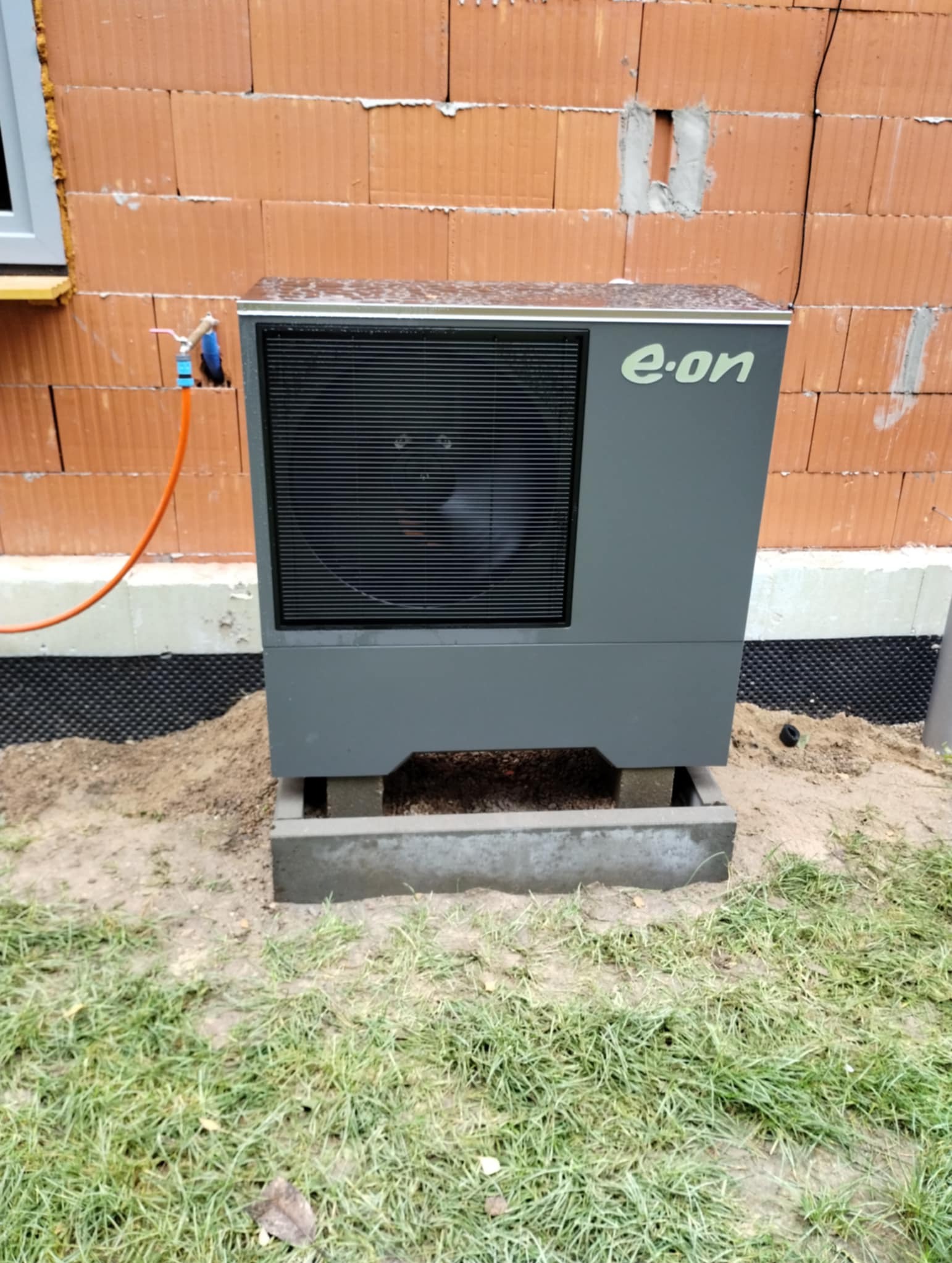Szara pompa ciepła marki E.ON zainstalowana na betonowych bloczkach przy ścianie budynku z czerwonej cegły, widoczny fragment trawnika i wężyk doprowadzający wodę.
