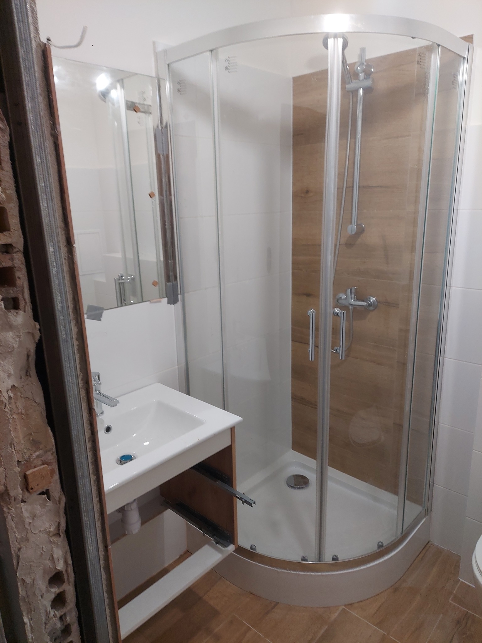 Niedokończona łazienka z kabiną prysznicową z drewnopodobnym panelem i umywalką z otwartą szafką, widoczne niedokończone ściany i elementy instalacji.