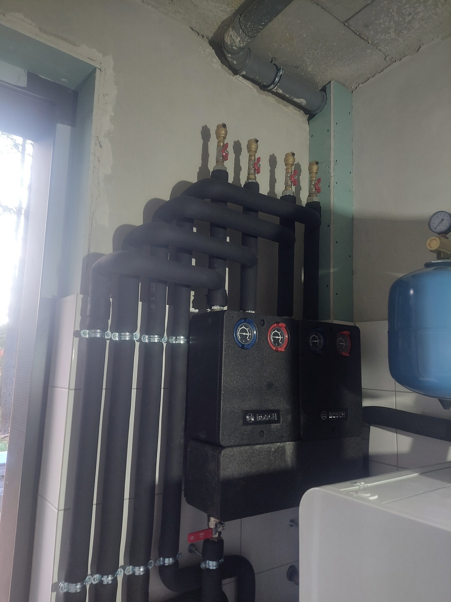 Instalacja grzewcza z czarnymi rurami w izolacji, manometrem, zaworami i modułem sterującym Bosch, widoczna przy ścianie w trakcie montażu.