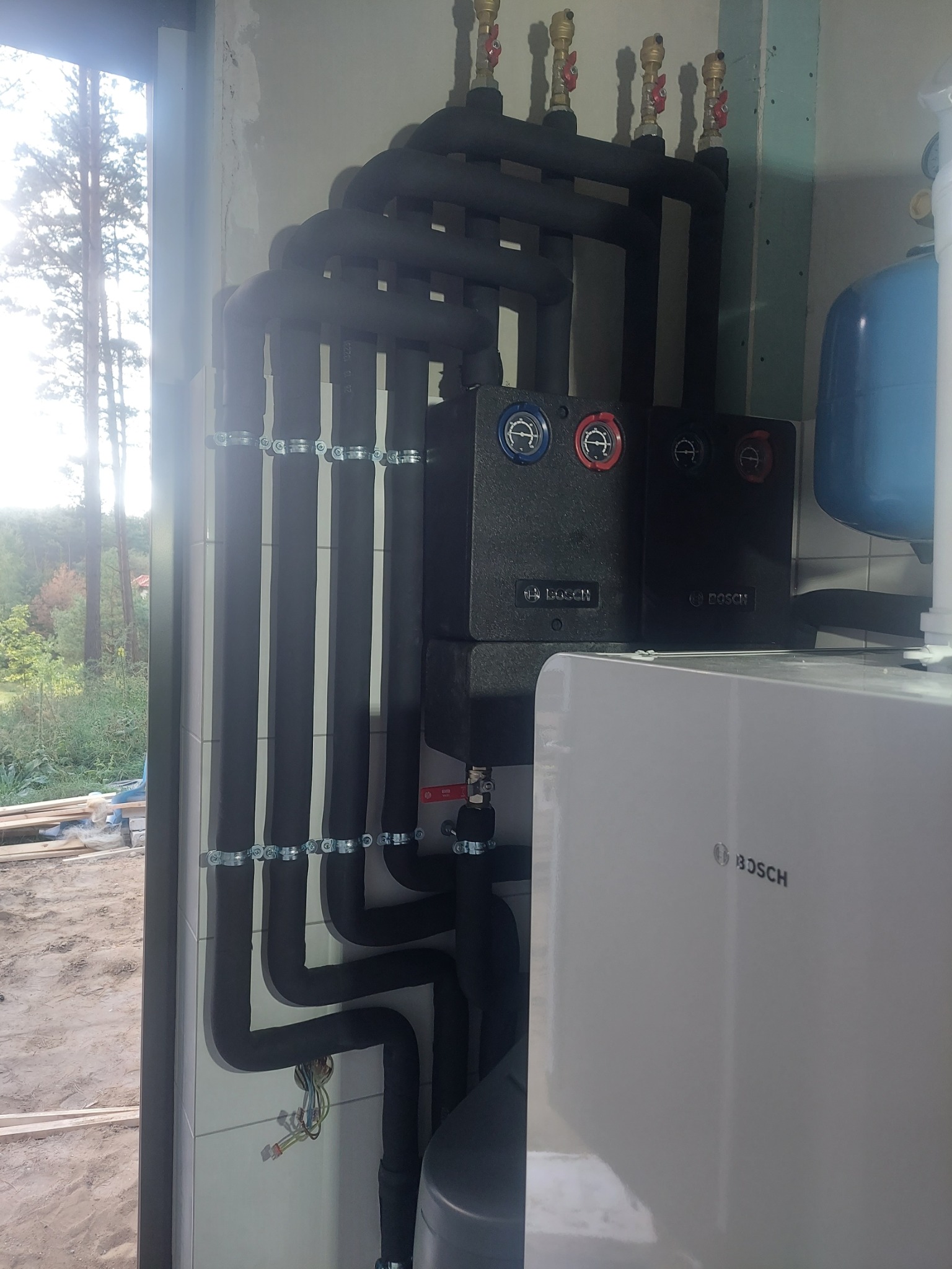 Nowa instalacja grzewcza z piecem gazowym marki Bosch, rurami w izolacji termicznej i osprzętem, zamontowana w surowym wnętrzu budynku w stanie deweloperskim, z widokiem na zewnątrz przez okno.