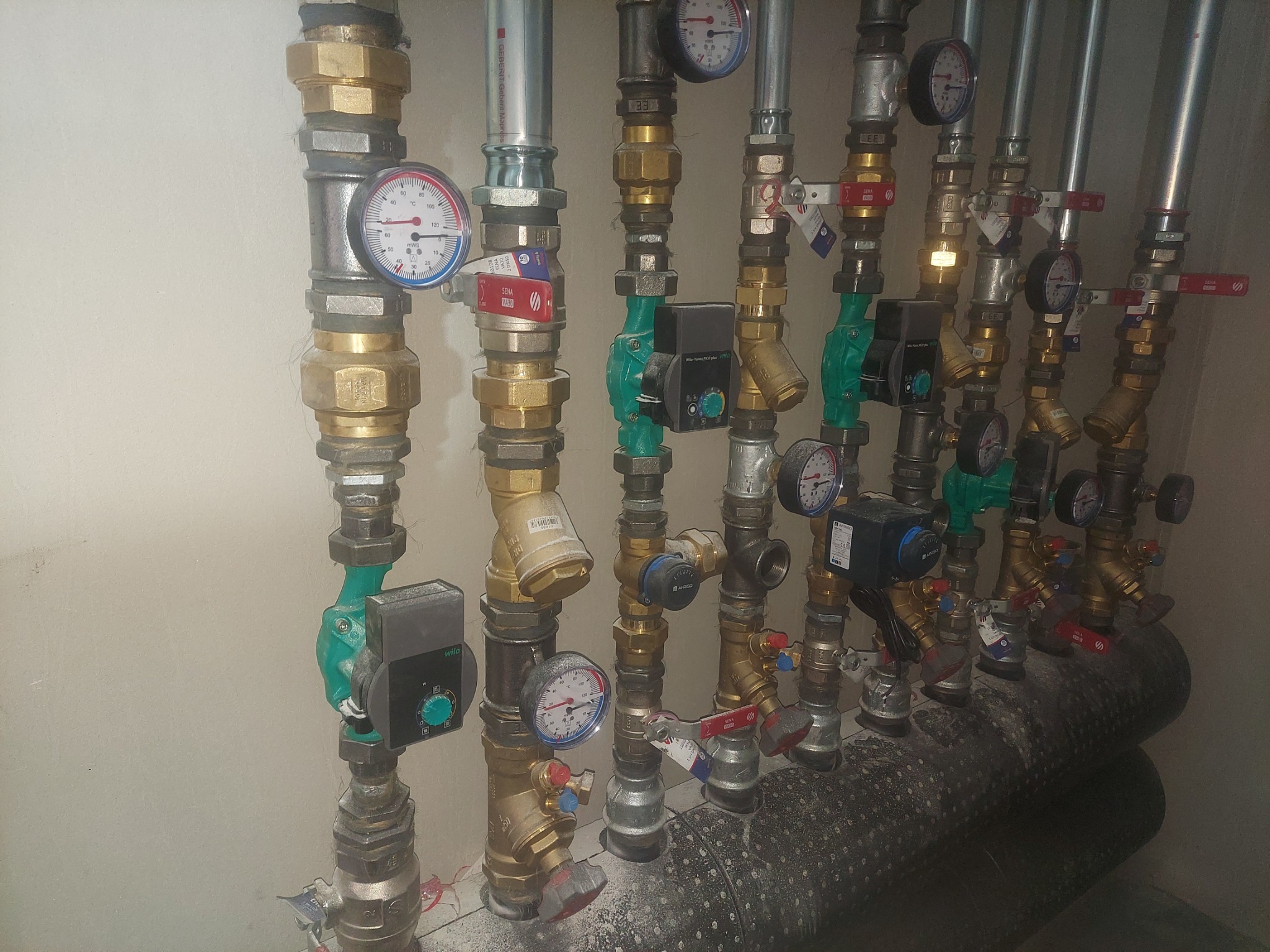 Rzędy pionowych rur instalacji grzewczej z manometrami, zaworami i pompami obiegowymi, widoczne izolowane poziome rury, przy ścianie, z bliska.