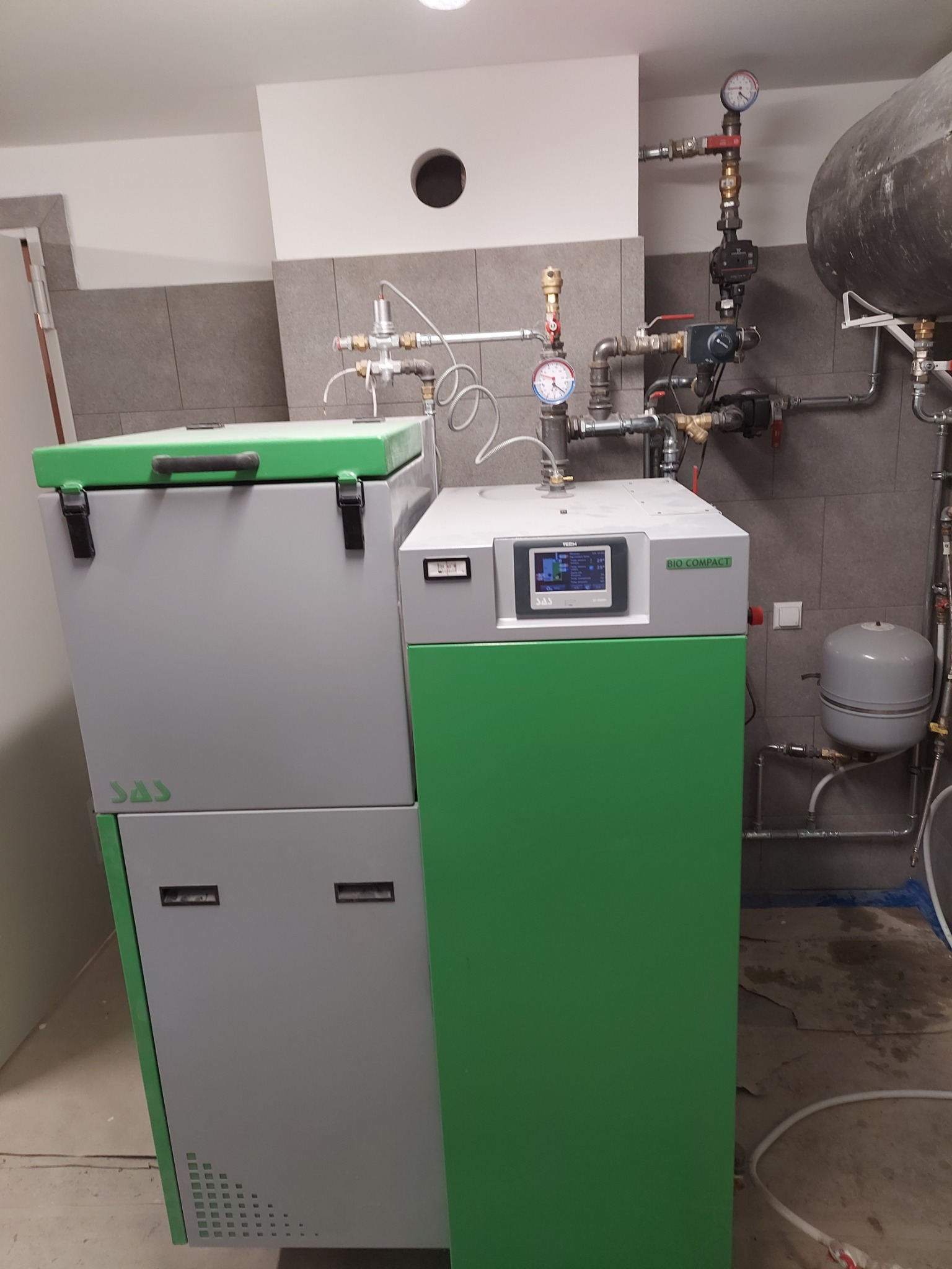 Nowoczesny piec CO Bio Compact SAS w kotłowni z szarymi płytkami, widoczne przyłącza hydrauliczne i zbiornik wyrównawczy.