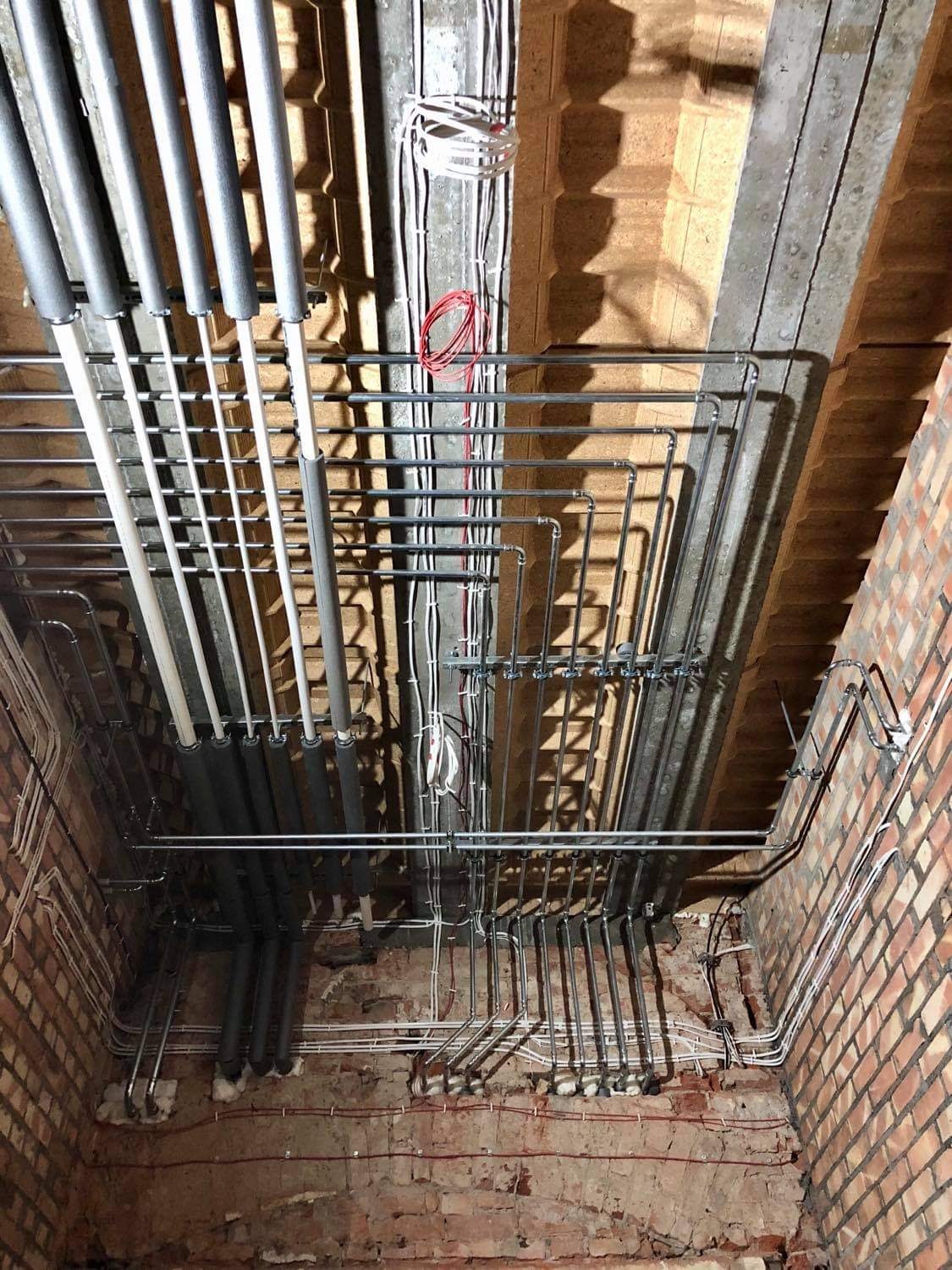 Widok instalacji grzewczej i elektrycznej pod stropem, z rurami w izolacji, przewodami oraz elementami mocującymi na surowej, ceglanej ścianie.