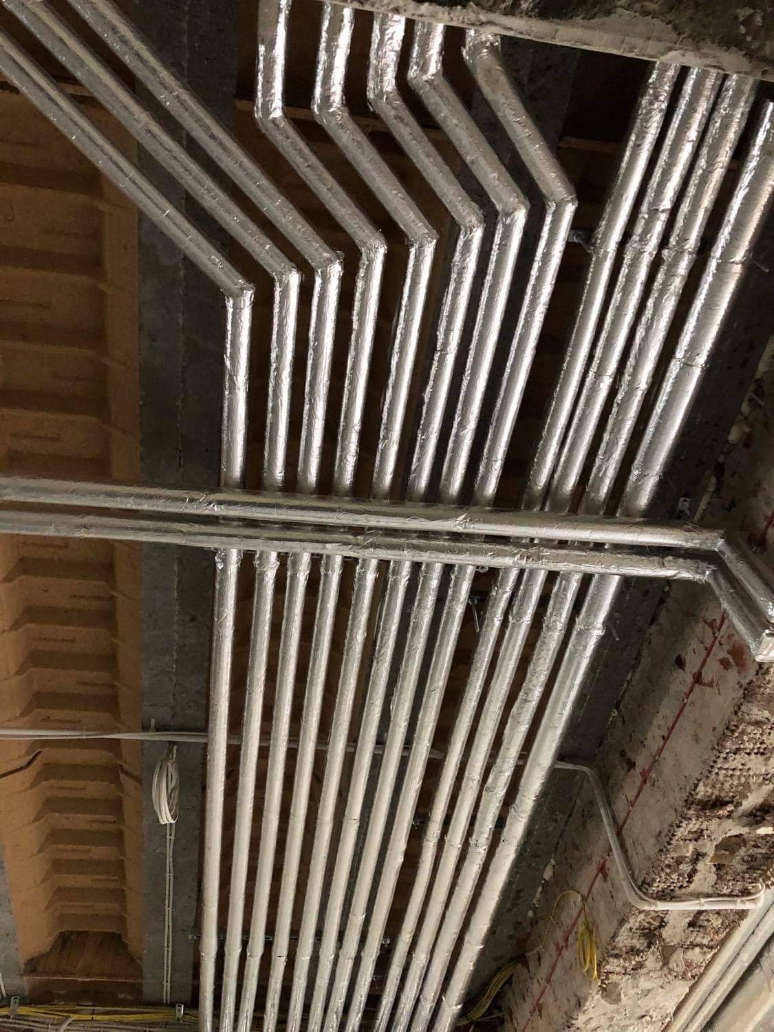 Instalacja rur grzewczych, zabezpieczonych srebrną izolacją, pod stropem w surowym stanie wykończenia, widoczne elementy podwieszenia i fragmenty instalacji elektrycznej.
