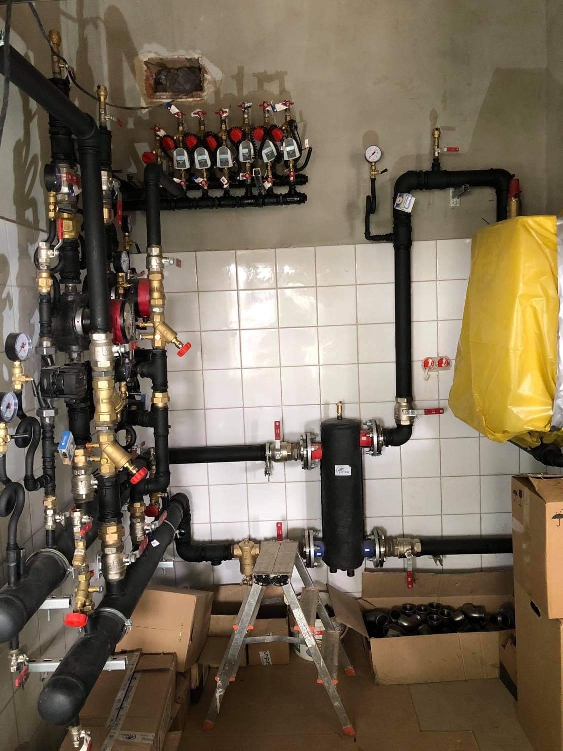 Rozbudowana instalacja hydrauliczna i grzewcza z licznymi rurami, zaworami i manometrami, zamontowana na ścianie wyłożonej białymi płytkami, z drabinką i kartonami na podłodze.