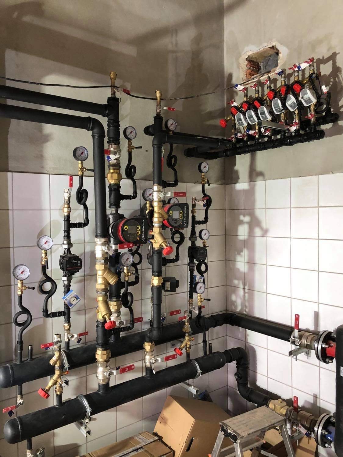 Złożona instalacja grzewcza z czarnych rur, pompami obiegowymi, manometrami i rozdzielaczem na ścianie z białych płytek, widoczna drabinka i kartonowe pudła.