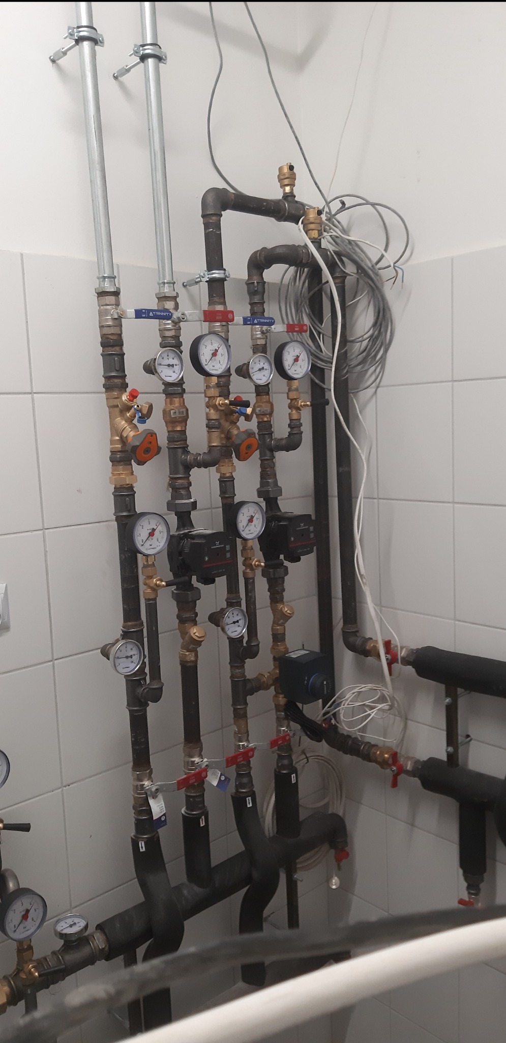 Rozbudowana instalacja hydrauliczna z rur miedzianych i stalowych, manometrami, zaworami i pompami obiegowymi w pomieszczeniu z białymi kafelkami.