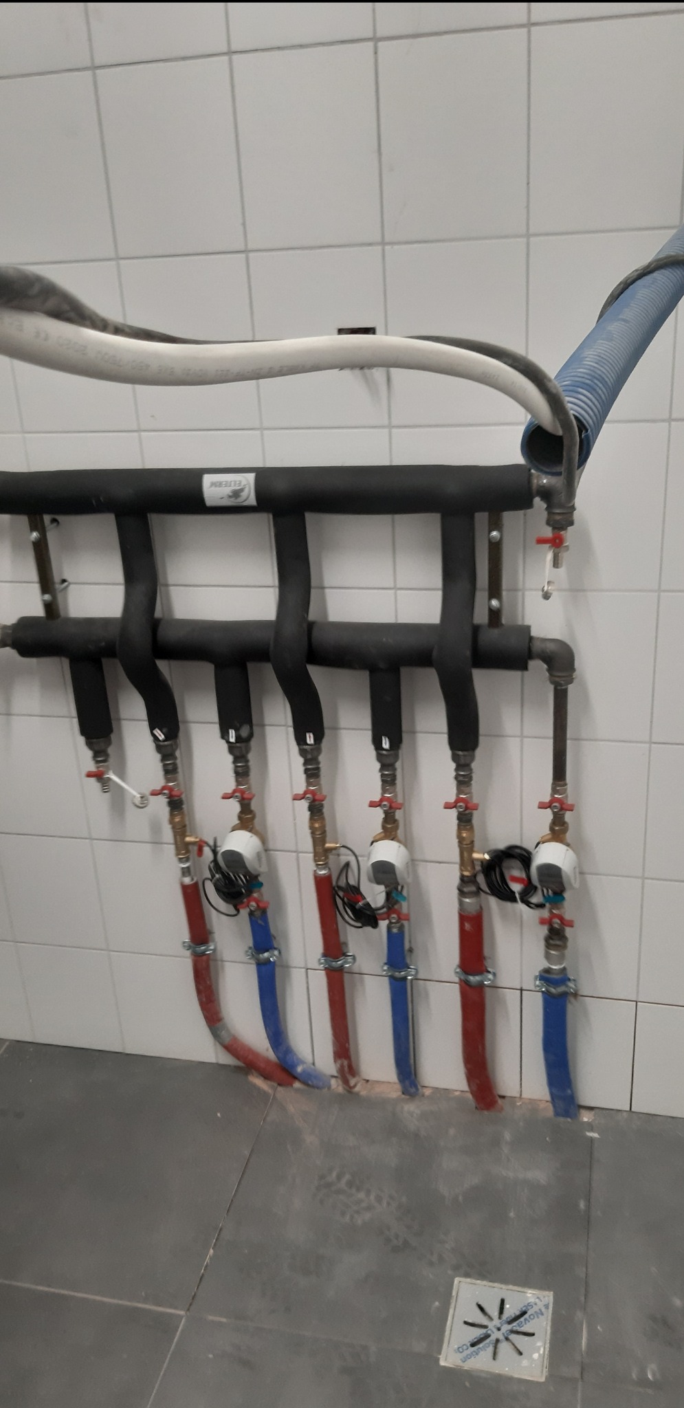 Rozdzielacz instalacji grzewczej z rurami w kolorach czerwonym i niebieskim, zaworami i licznikami, zamontowany na ścianie z białych płytek w pomieszczeniu technicznym, z widoczną kratką ściekową...