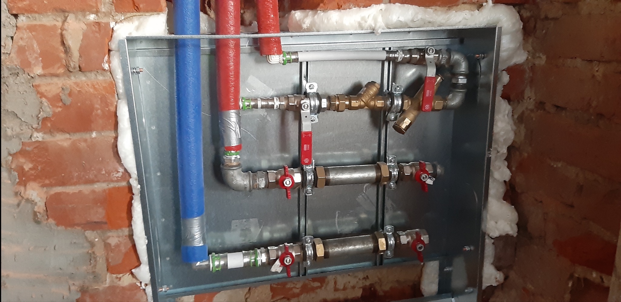 Rozdzielacz instalacji wodnej w metalowej obudowie, z rurami w izolacji czerwonej i niebieskiej, zamontowany na tle ceglanej ściany z pianką uszczelniającą.