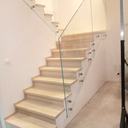 Phu eurokrys Krystian Kozioł - Jasne, drewniane stopnie schodów z przezroczystą szklaną balustradą mocowaną metalowymi uchwytami w nowoczesnym wnętrzu.