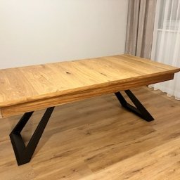 Phu eurokrys Krystian Kozioł - Rozkładany stół z blatem z jasnego drewna i czarnymi, geometrycznymi nogami, ustawiony na podłodze imitującej drewno, z zasłonami w tle.