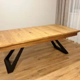 Rozkładany stół z blatem z jasnego drewna i czarnymi, geometrycznymi nogami, ustawiony na podłodze imitującej drewno, z zasłonami w tle.