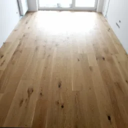 Ułożona podłoga z desek dębowych z widocznymi sękami i naturalnym usłojeniem, jasne wnętrze z dużym oknem w tle, widoczny grzejnik zabezpieczony folią.