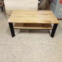 Phu eurokrys Krystian Kozioł - Stolik kawowy z jasnego drewna dębowego z półką i czarnymi, metalowymi nogami, na tle warsztatu z maszynami do obróbki drewna.