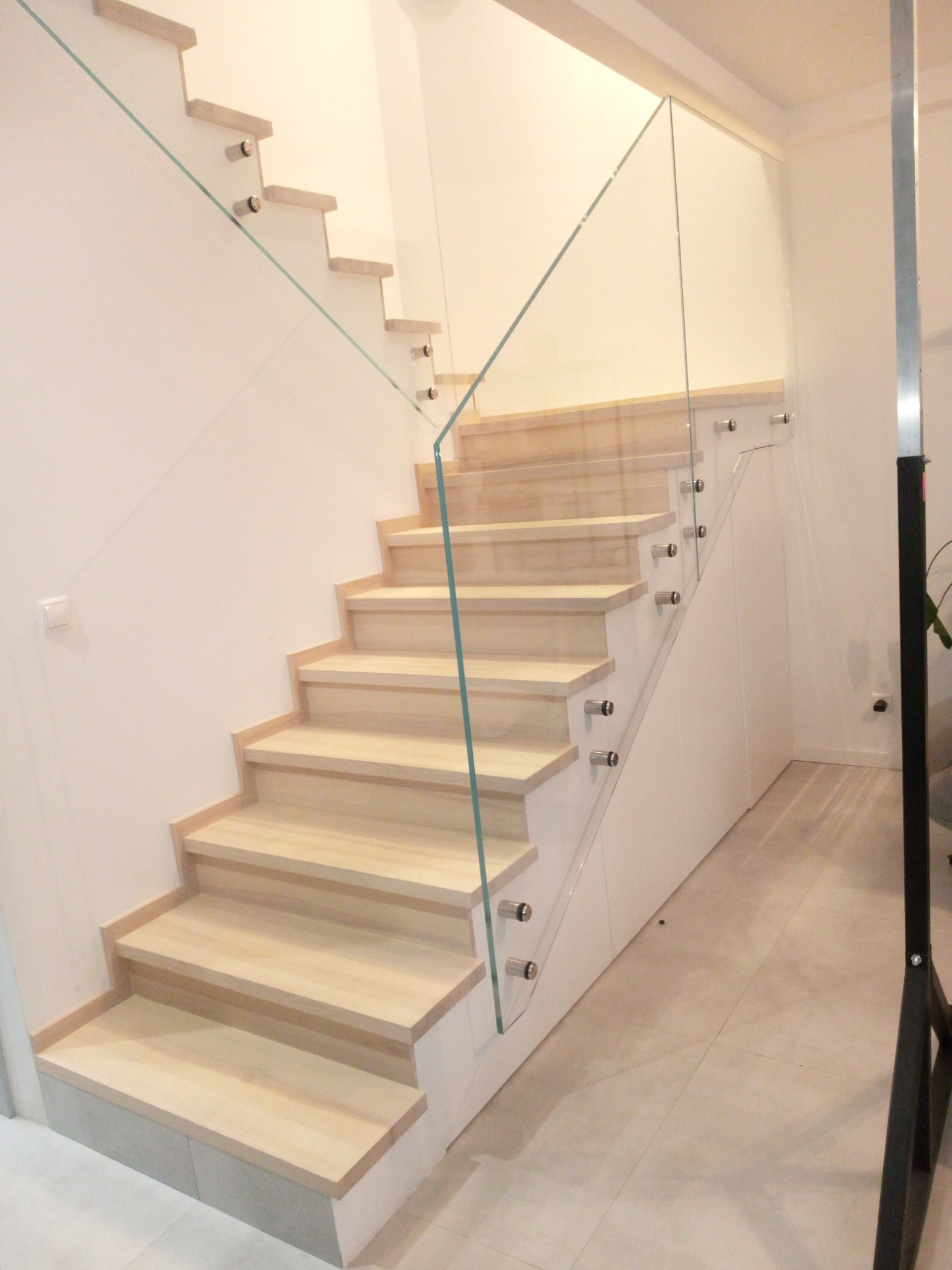 Jasne, drewniane stopnie schodów z przezroczystą szklaną balustradą mocowaną metalowymi uchwytami w nowoczesnym wnętrzu.
