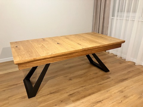 Rozkładany stół z blatem z jasnego drewna i czarnymi, geometrycznymi nogami, ustawiony na podłodze imitującej drewno, z zasłonami w tle.