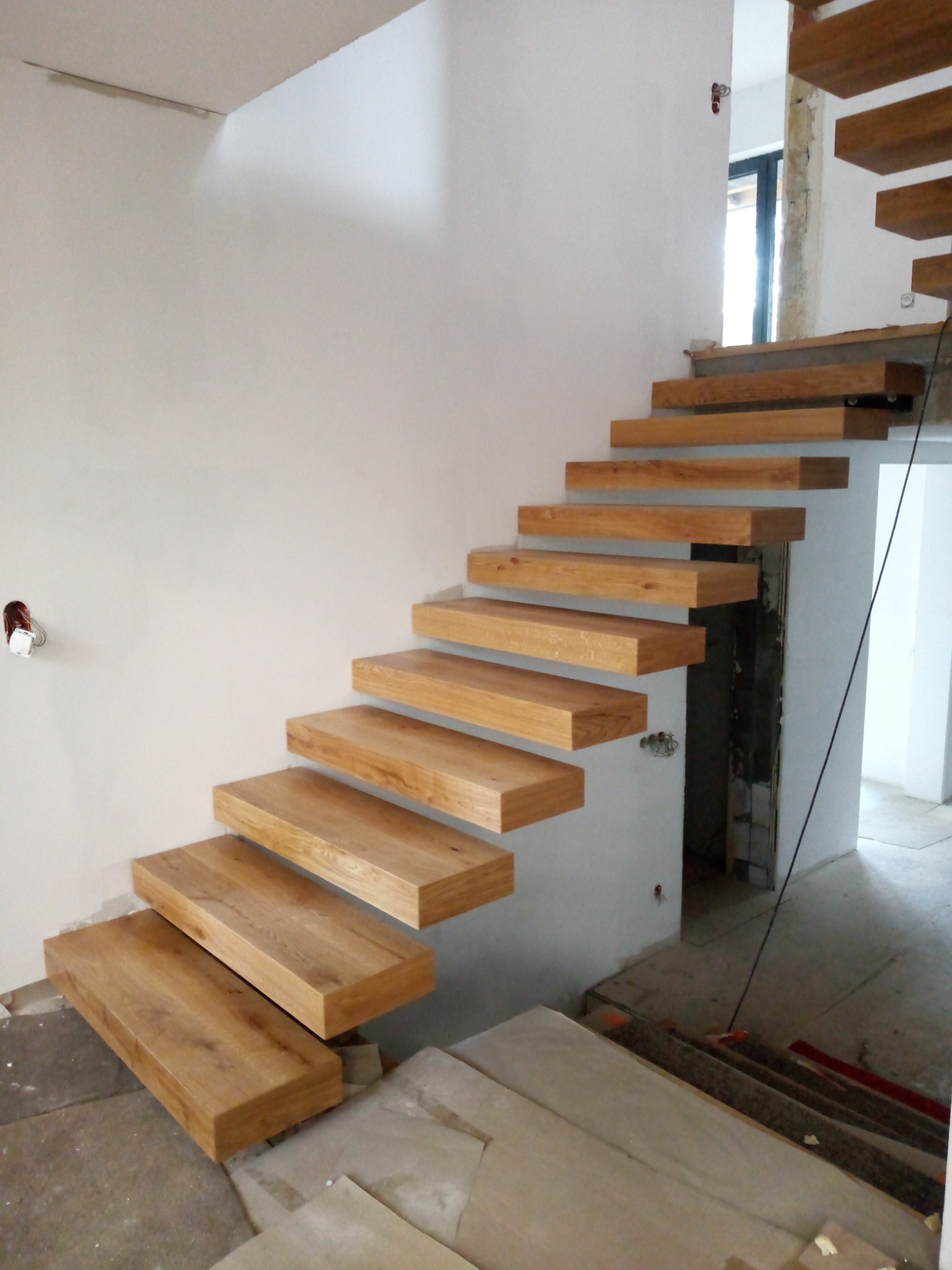 Ażurowe schody z jasnego drewna zamontowane w surowym wnętrzu, widoczne niedokończone ściany i podłoga w trakcie wykańczania.