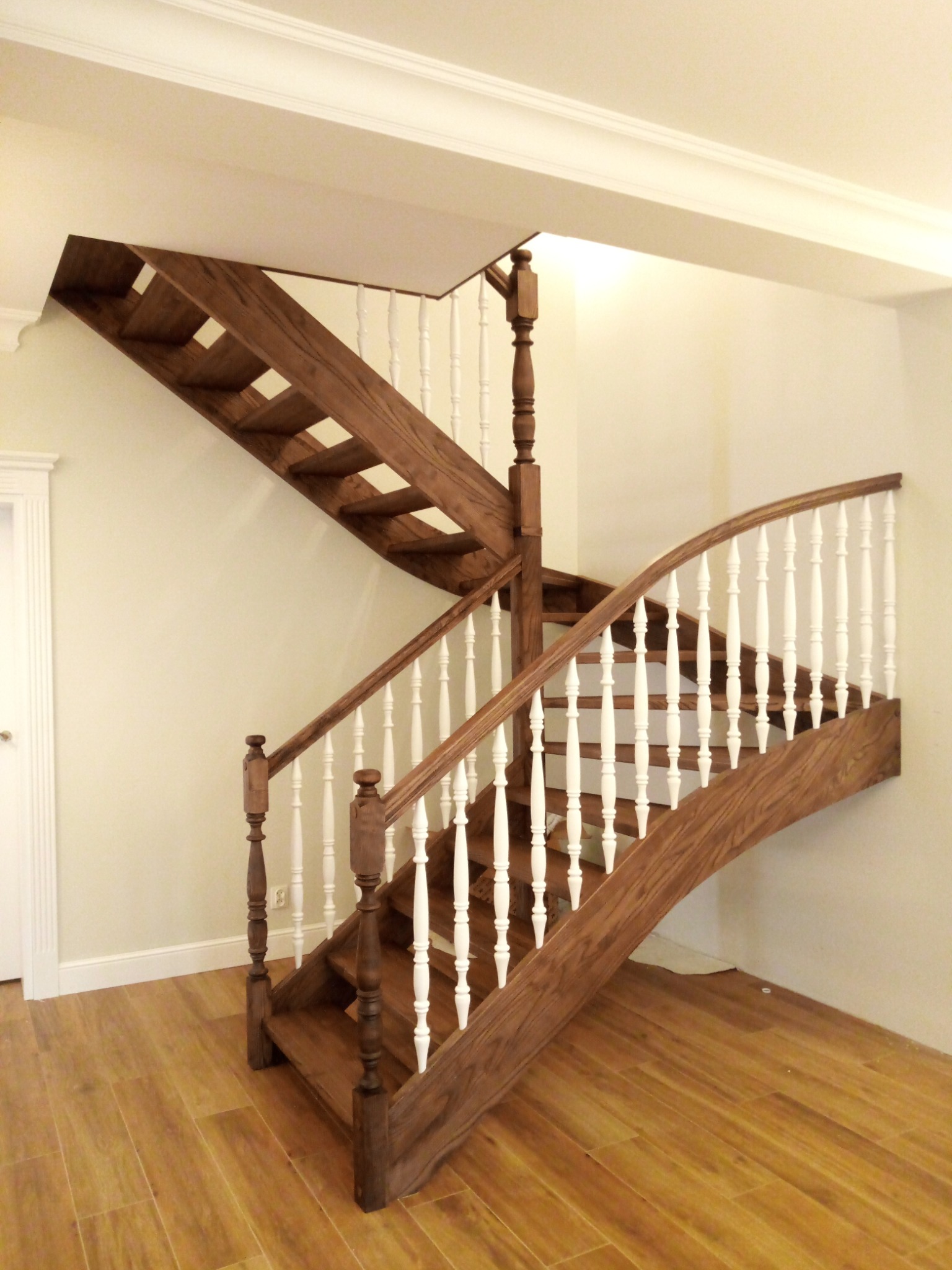 Dwubiegowe schody z drewna o ciemnym wybarwieniu, z białymi tralkami i zakrętem, na podłodze z paneli imitujących drewno.