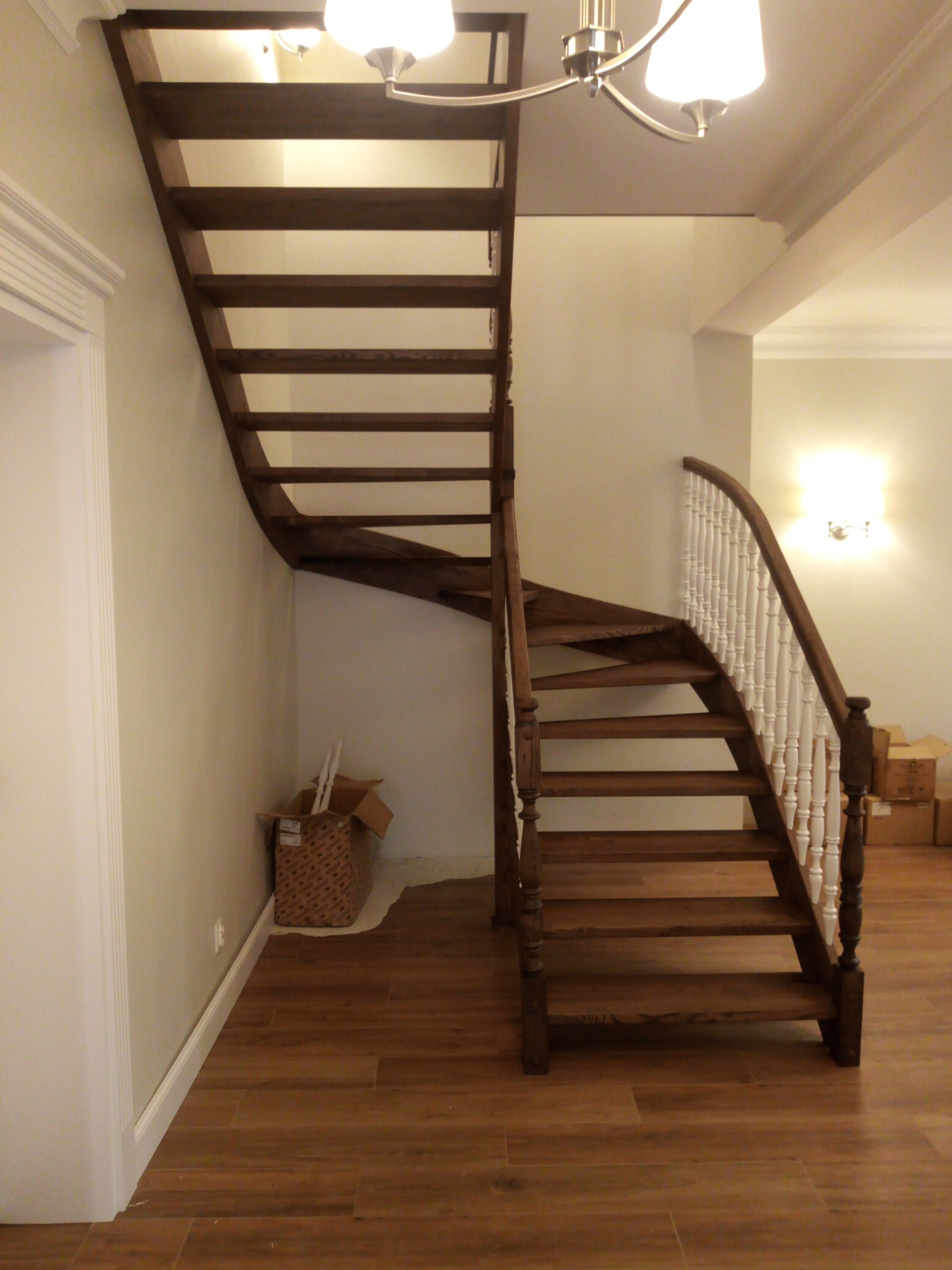 Drewniane schody z białą balustradą w jasnym wnętrzu, widoczne kartony i elementy wykończeniowe na podłodze.