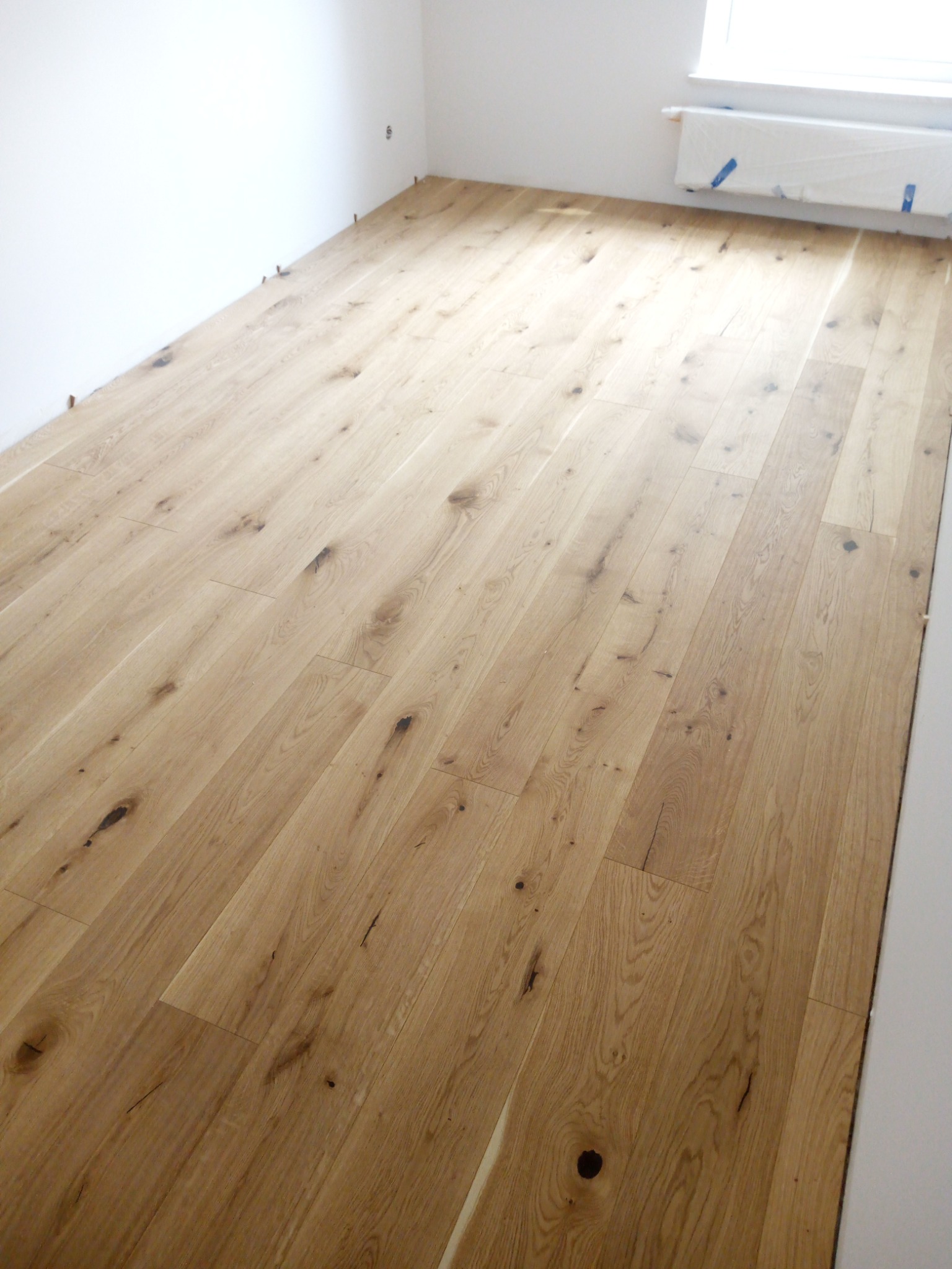 Ułożona podłoga z jasnych, drewnianych desek z widocznymi sękami i usłojeniami, przy białej ścianie z klinami dystansowymi i zakrytym grzejnikiem.