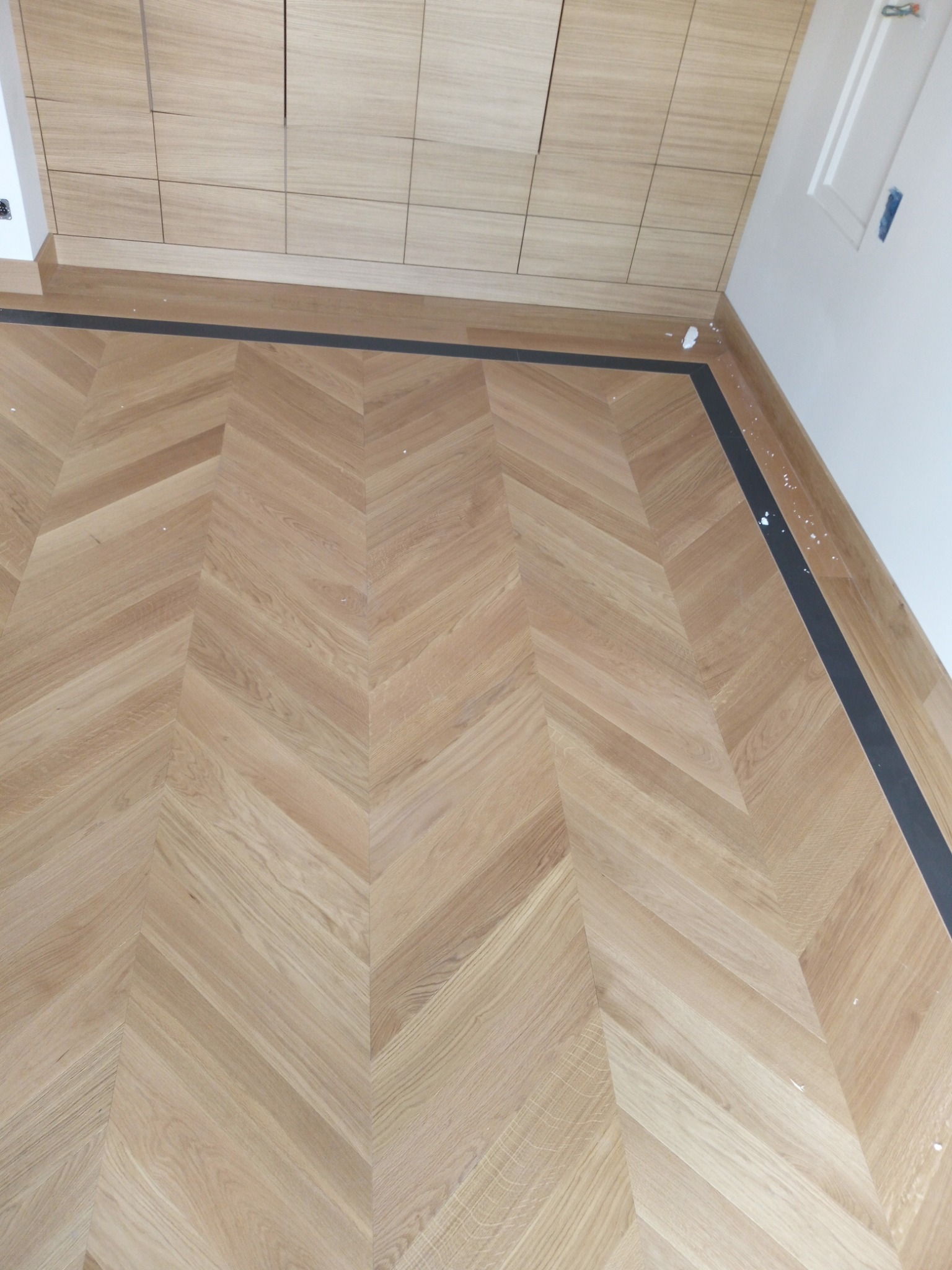 Ułożona w jodełkę podłoga z jasnego drewna w Hajnówce, z czarną listwą przypodłogową i fragmentem zabudowy meblowej u góry kadru.