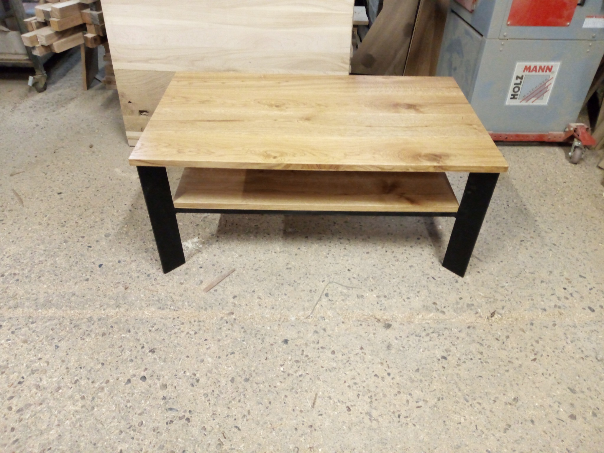 Stolik kawowy z jasnego drewna dębowego z półką i czarnymi, metalowymi nogami, na tle warsztatu z maszynami do obróbki drewna.