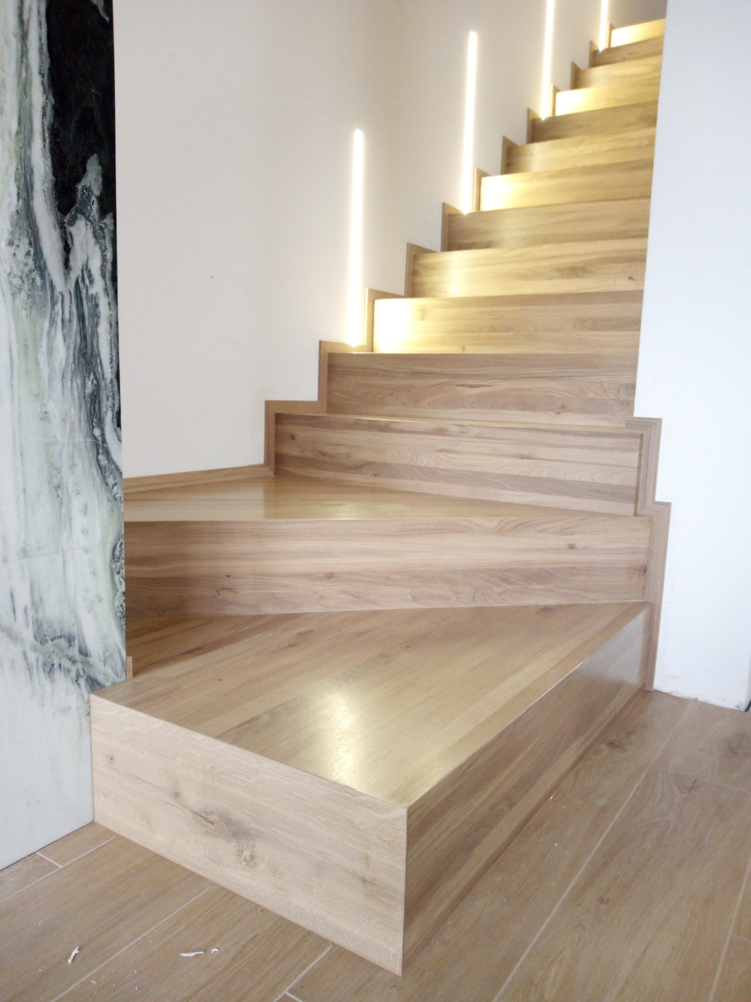 Nowoczesne schody z jasnego drewna z podświetleniem LED wzdłuż stopni, marmurowa okładzina ściany po lewej stronie, widoczne usłojenie drewna.