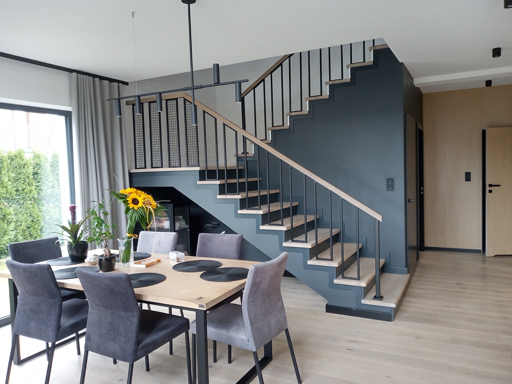 Nowoczesne schody z metalową balustradą i drewnianymi stopniami w minimalistycznym wnętrzu z jadalnią, oświetlone wiszącą lampą.