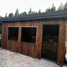 Drewniana altana z ciemnymi szybami i płaskim dachem, otoczona młodymi drzewkami. Widoczne detale wykończenia i elementy otoczenia w naturalnym krajobrazie.