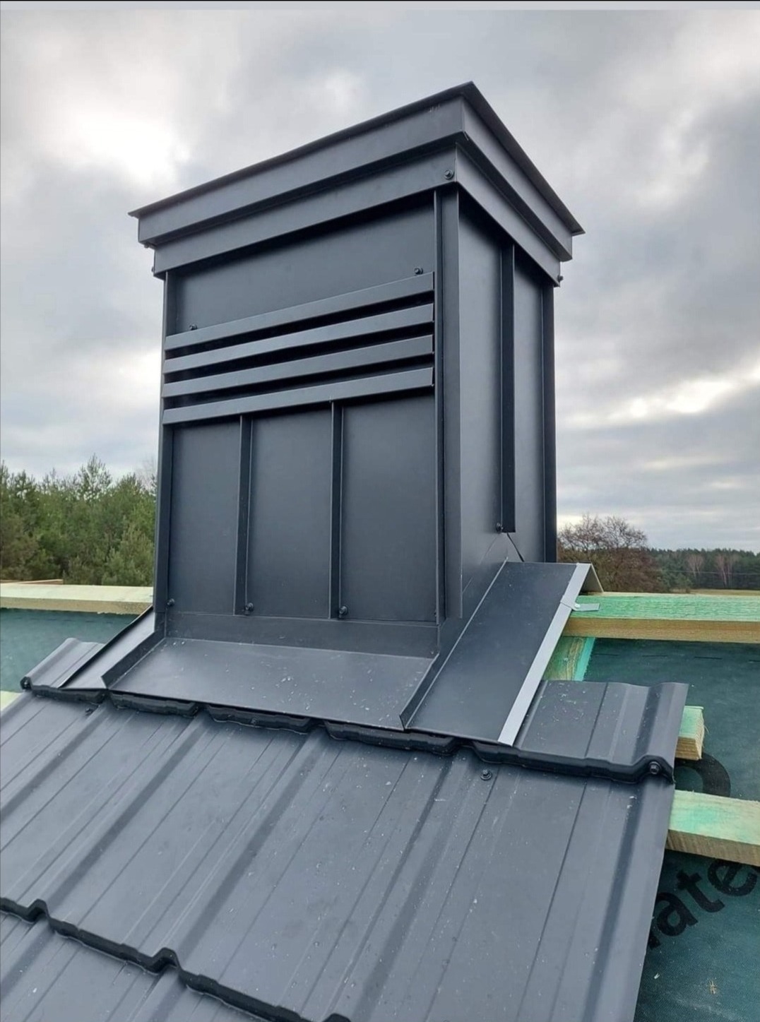 Szary komin wentylacyjny z blachy, zamontowany na dachu z blachodachówki w trakcie montażu, widoczne drewniane łaty i membrana dachowa.