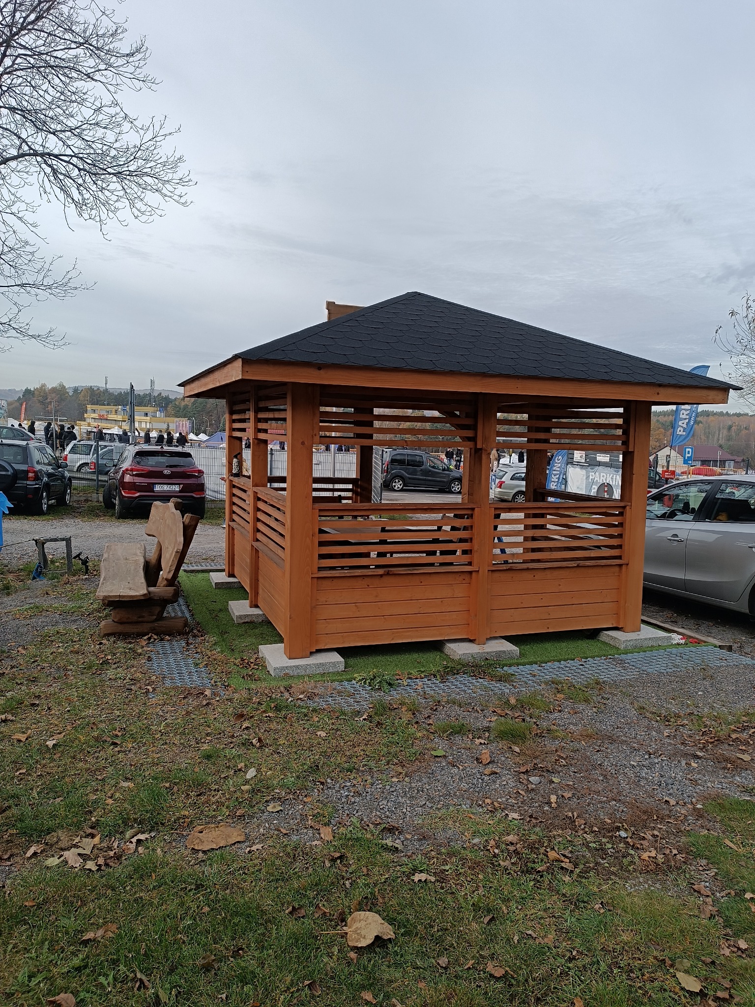 Drewniana altana z czarnym dachem bitumicznym, z ażurowymi ścianami, umieszczona na trawniku z kostki ażurowej, w tle parking i zabudowania.