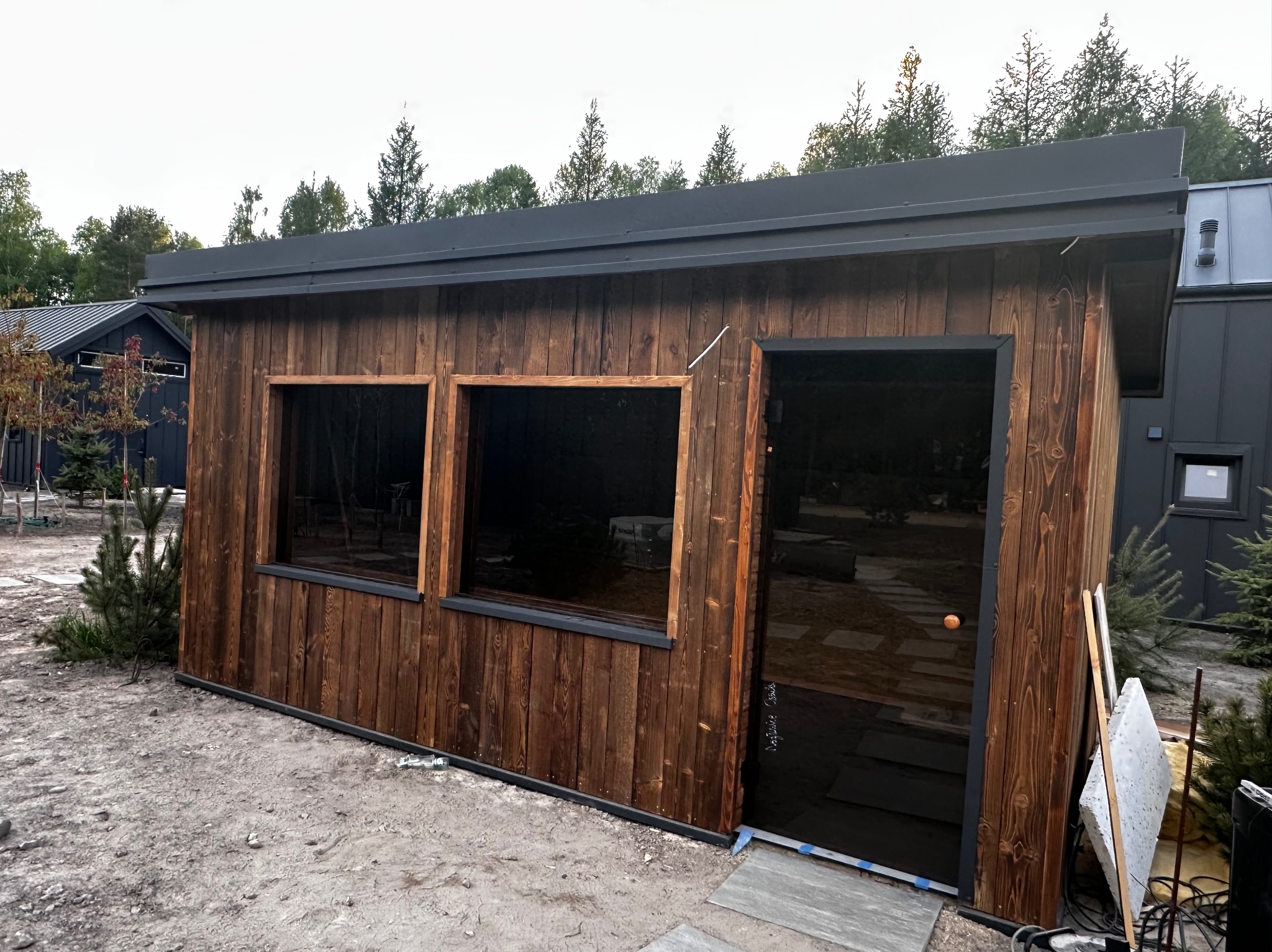 Drewniana altana z ciemnymi szybami i płaskim dachem, otoczona młodymi drzewkami. Widoczne detale wykończenia i elementy otoczenia w naturalnym krajobrazie.