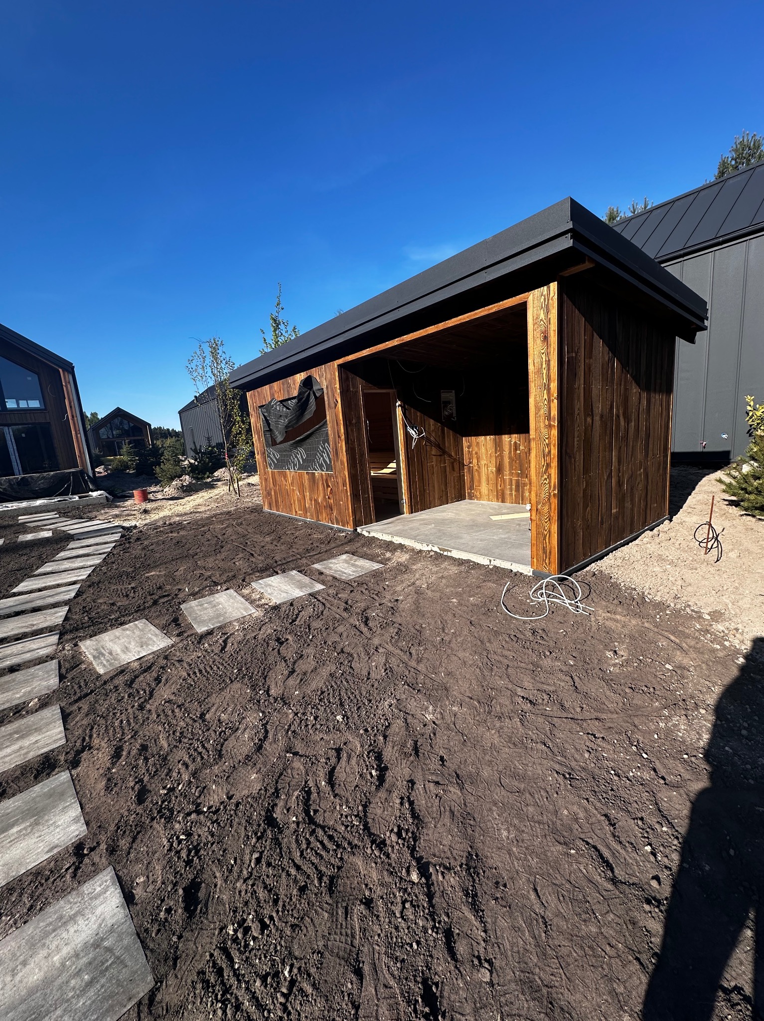 Nowoczesna, drewniana altana z płaskim, ciemnym dachem w trakcie budowy. Widoczne elementy konstrukcyjne i otoczenie z ułożonymi płytami chodnikowymi.