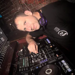 DJ z fioletowymi słuchawkami Pioneer na szyi, miksujący muzykę na profesjonalnym sprzęcie Pioneer DJ w dimly oświetlonym klubie z ceglaną ścianą w tle.