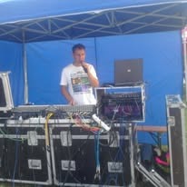 Mężczyzna z mikrofonem za konsoletą DJ-ską pod niebieskim namiotem, laptop w tle, sprzęt oświetleniowy widoczny.