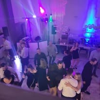 Tańczący goście na sali weselnej oświetlonej fioletowymi światłami, w tle widoczny sprzęt nagłośnieniowy i dekoracje.