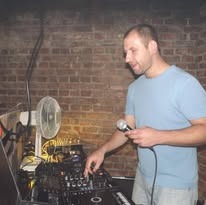 Mężczyzna w niebieskiej koszulce obsługuje sprzęt audio na tle ceglanej ściany, trzymając mikrofon w dłoni.