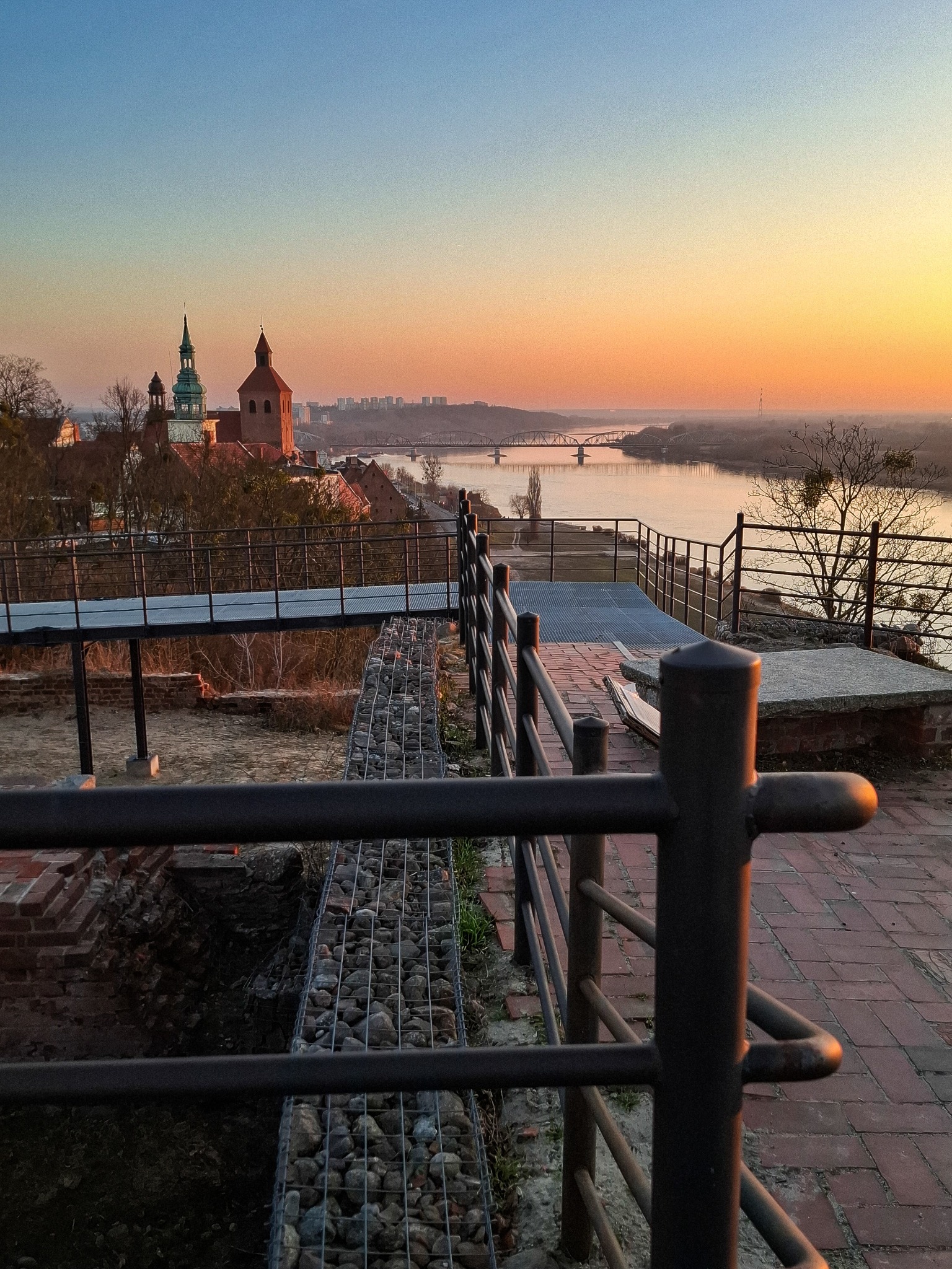 Panorama miasta nad rzeką o wschodzie słońca, widok z tarasu z metalową barierką, fragmenty murów obronnych i kosze gabionowe.